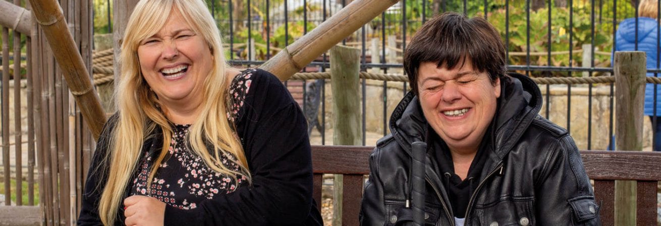 Two ladies from Ambient Support's Learning Disability services are laughing together, they are being supported on a day out. Supported living, short breaks, respite.