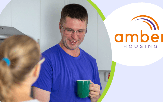 A man is holding a cup of tea while talking to his support worker, he is wearing a blue shirt. There is text next to the image which reads 'Amber Housing'.
