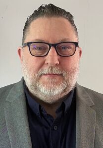 Photo of troy Henshall, CEO of amber housing, he is a white male wearing a grey suit with black rimmed glasses and a white short beard.